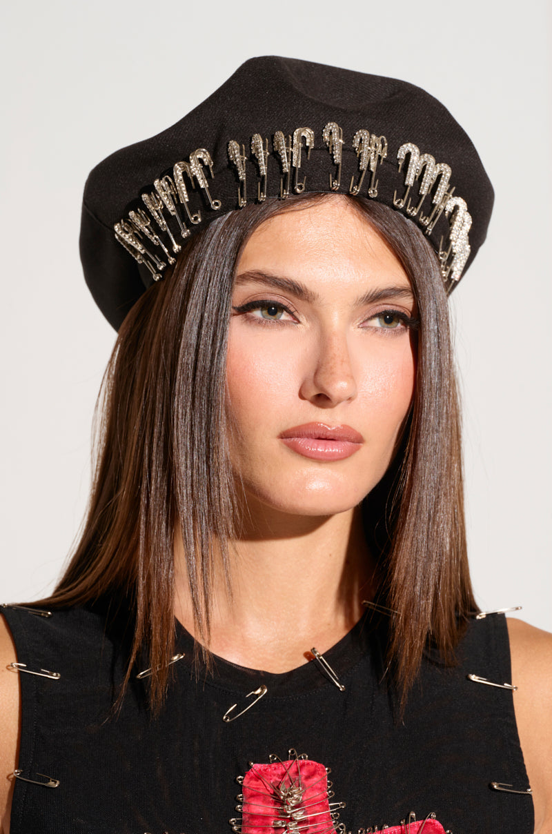 A woman with straight, brown hair wears the SAFETY FIRST BERET from AKIRA Label, accented with silver safety pins. She is also dressed in a coordinating black top featuring similar safety pin embellishments. She gazes forward with a neutral expression against a plain background. 0
