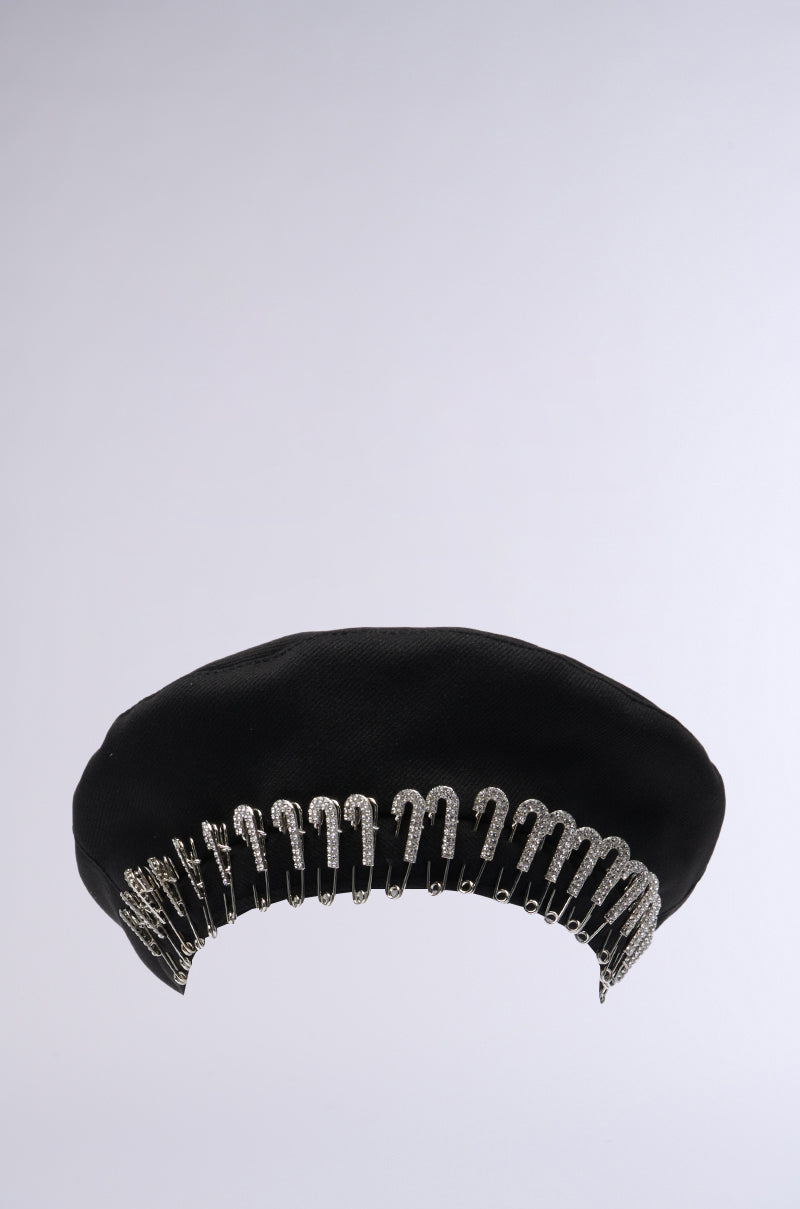 The SAFETY FIRST BERET from the AKIRA Label features a series of evenly spaced silver safety pins around the rim, creating a unique and decorative pattern. The plain, light gray background accentuates the beret's distinctive style. 2