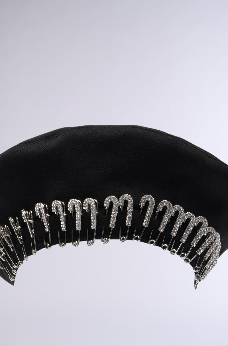 The SAFETY FIRST BERET by AKIRA Label is a stylish black beret adorned with a row of silver safety pins evenly spaced along the edge, adding a striking metallic accent to the hat's fabric.