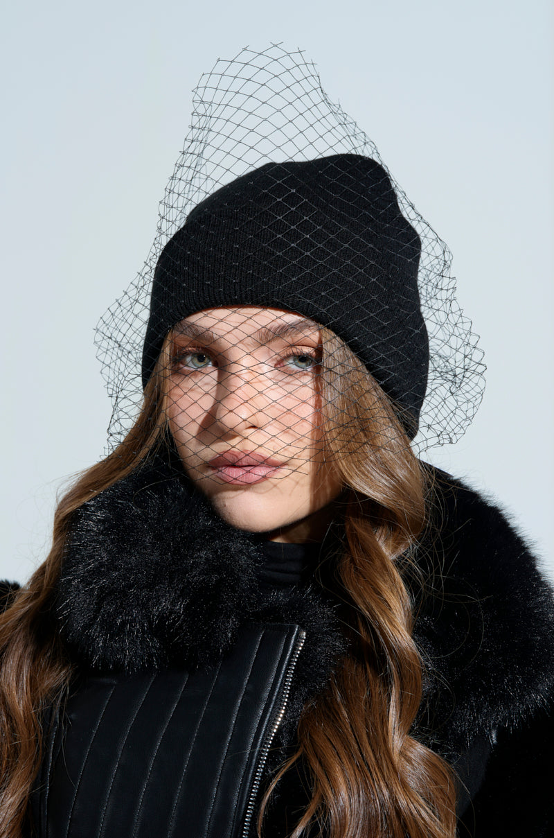 A person with long hair is wearing a ROCKY MESH VEIL BEANIE and a black fur-collared coat, posing against a light background.