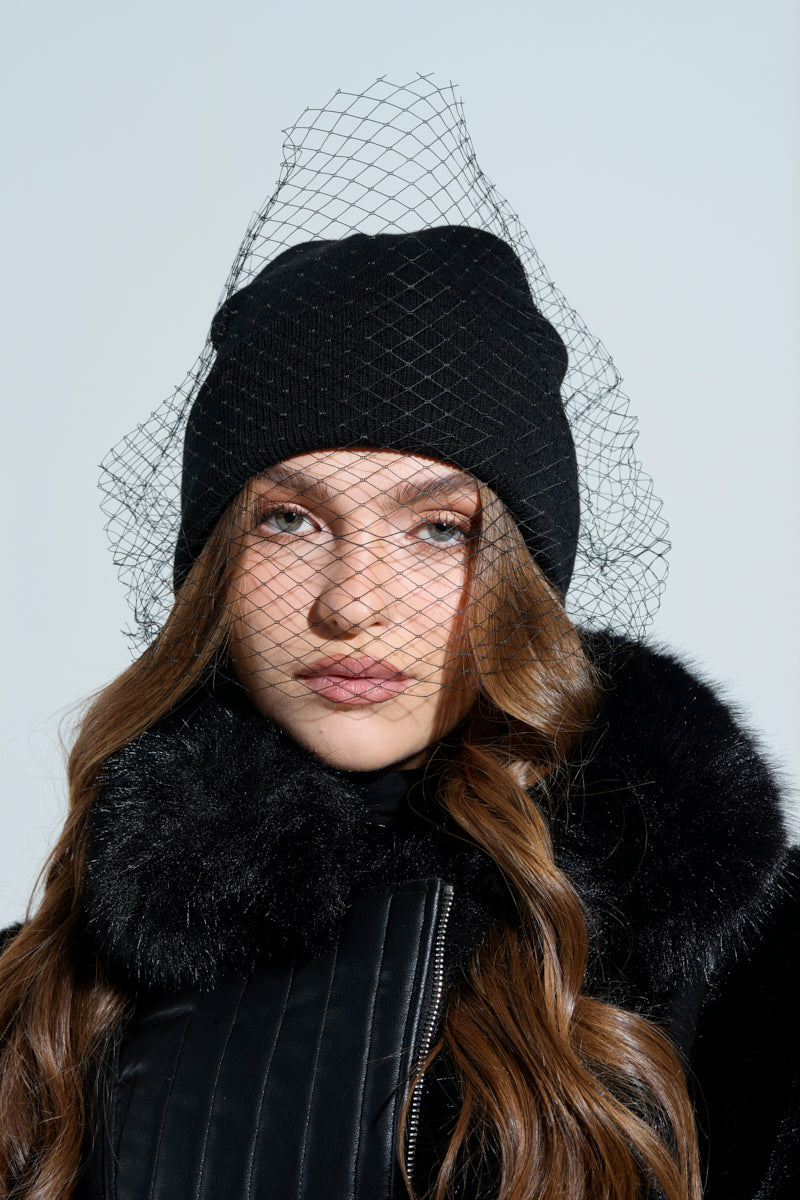 Wearing a ROCKY MESH VEIL BEANIE, a person poses for a portrait in a black coat with a fur collar. Their long hair falls in loose waves over their shoulders against a neutral light-toned background.