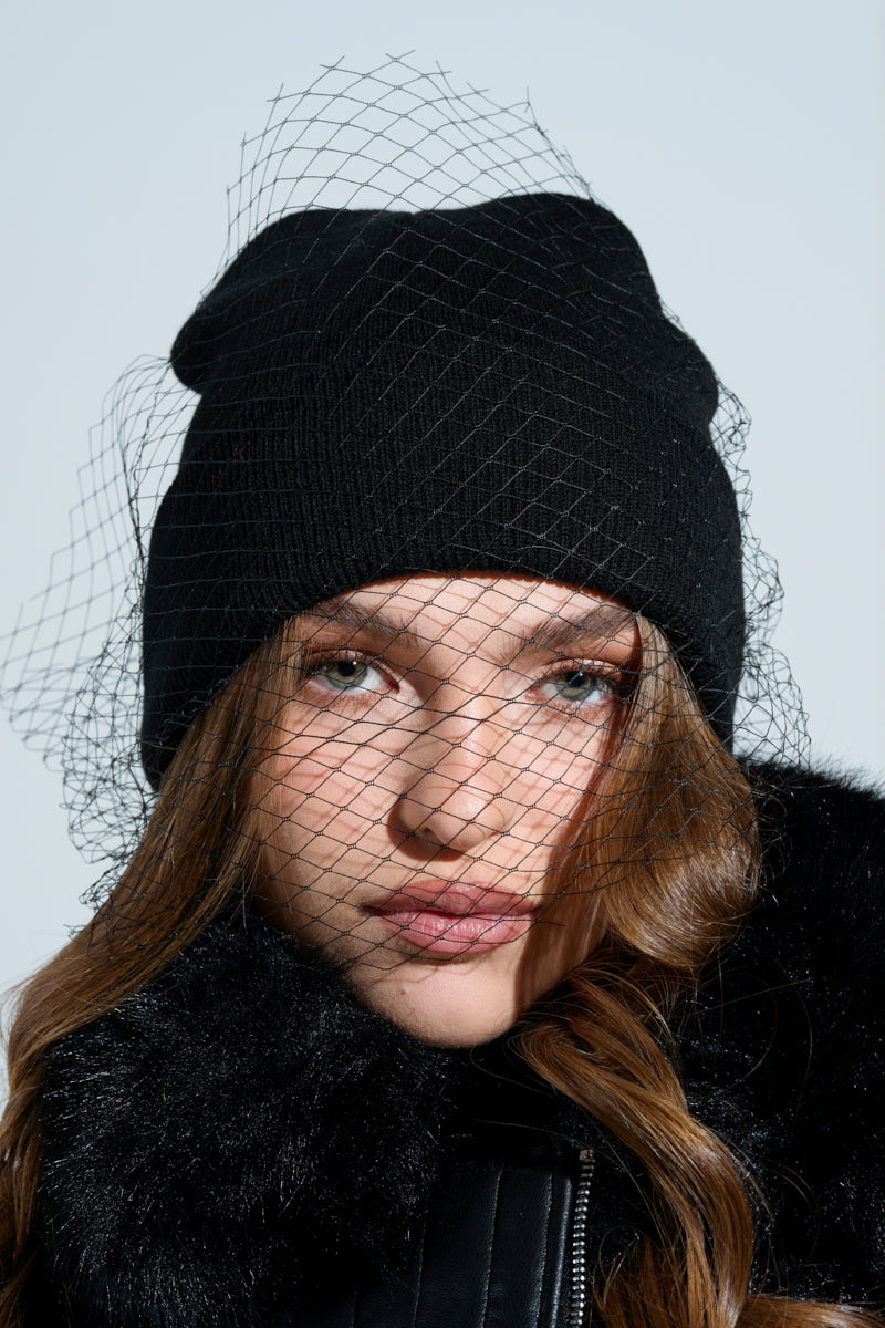 A person wearing the ROCKY MESH VEIL BEANIE and a black fur-collared coat looks directly at the camera against a light, neutral background.