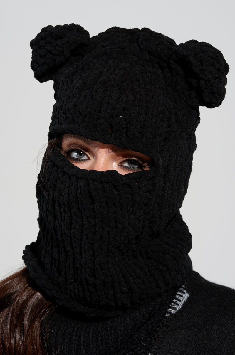 A person wears the HEARD YOU CROCHET KNIT BALACLAVA in black, featuring bear ears with only their eyes visible. The thick, textured fabric envelops most of their face and neck. This distinct winter accessory stands out against the plain, light-colored background.