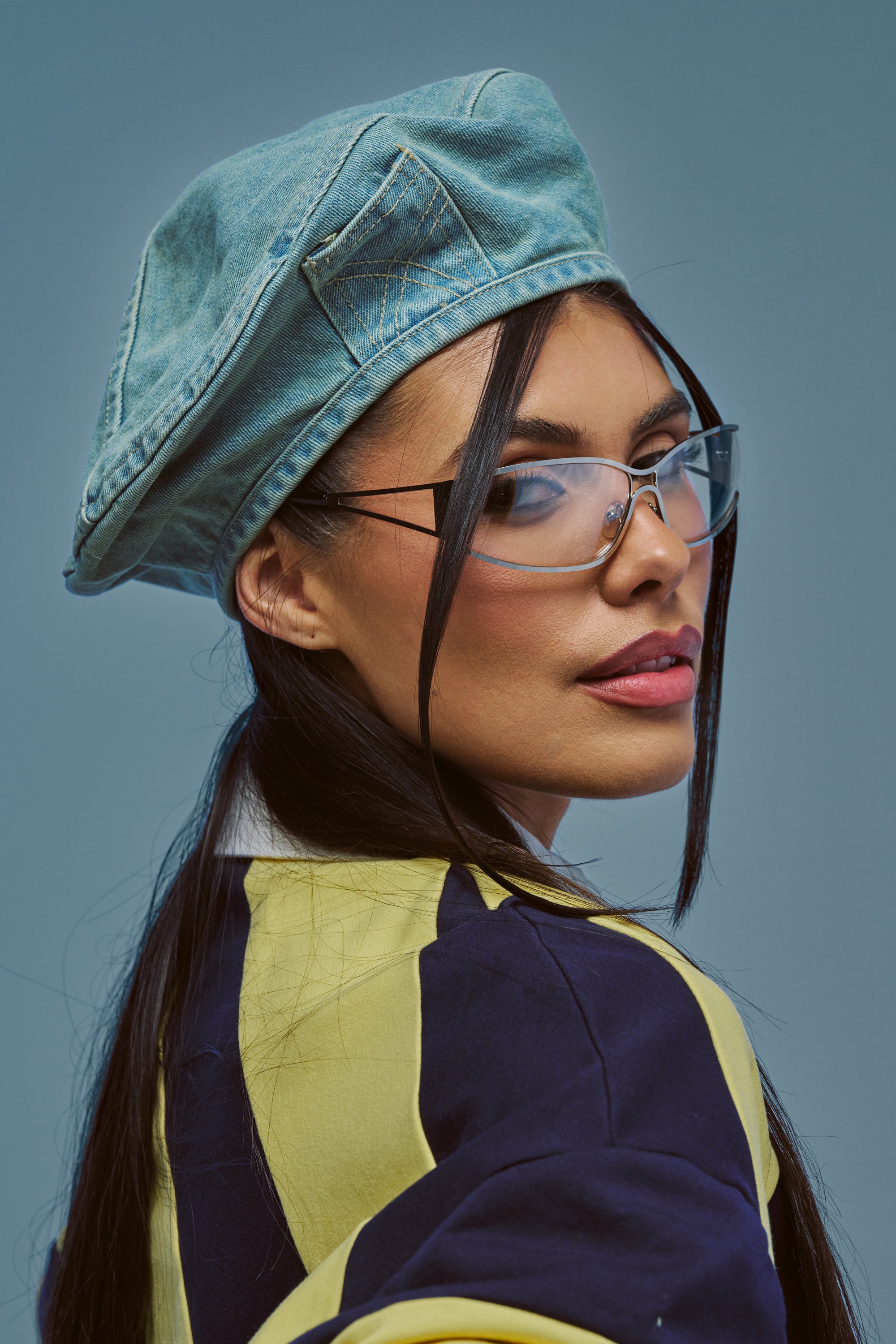 Wearing the IN MY POCKET DENIM BERET in a light wash, clear glasses, and a yellow-and-black jacket, the woman channels effortless streetwear style as she glances over her shoulder against a plain blue background.