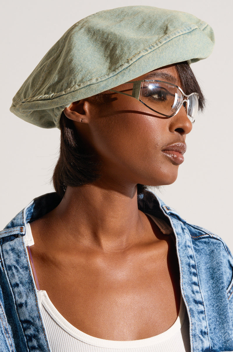 A person wearing the IN MY POCKET DENIM BERET by TOROS ACCESSORIES and transparent glasses is depicted in profile. They have a short haircut and are dressed in a white top under a light wash denim jacket. The neutral background highlights their stylish, modern look, accentuated by the perfect autumn accessory.