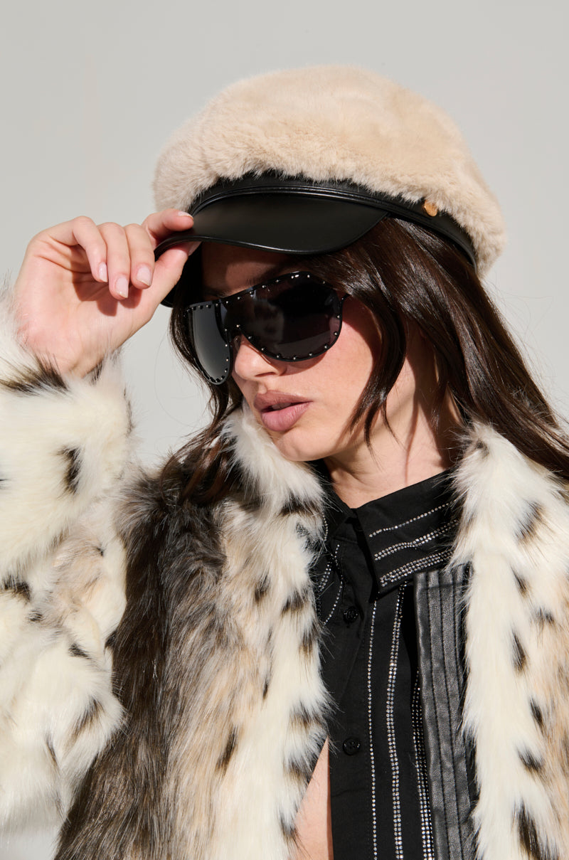 A person wearing a faux fur coat and the IN THE CITY FUR BRIM HAT adjusts their sunglasses. The outfit is chic, blending a mix of textures and dark tones, set against a plain background.