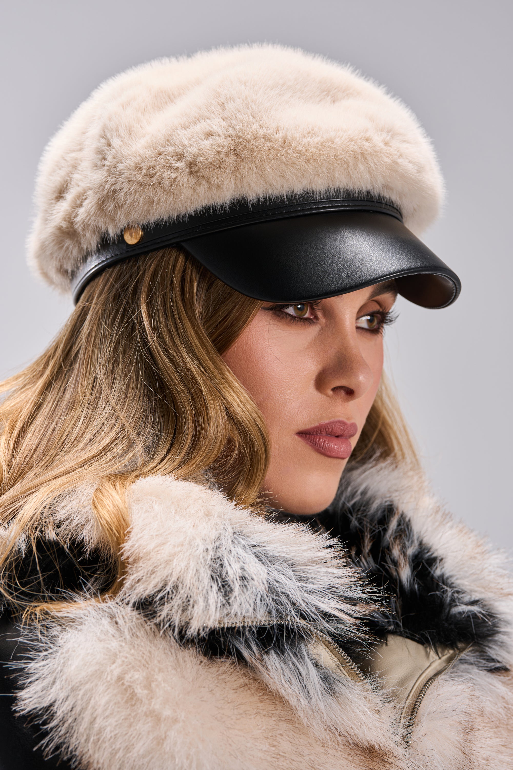 A woman wears the IN THE CITY FUR BRIM HAT—a beige faux fur fisherman cap with a black leather brim—paired with a matching fur-collared coat, gazing to the side against a neutral background.