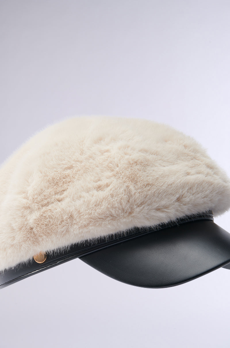 Introducing the IN THE CITY FUR BRIM HAT: a chic fisherman-style cap that boasts a plush beige faux fur top, complemented by a sleek black leather brim adorned with a gold button accent on the side. This exquisite piece stands out against a soft, neutral gray background, emphasizing its luxurious texture.