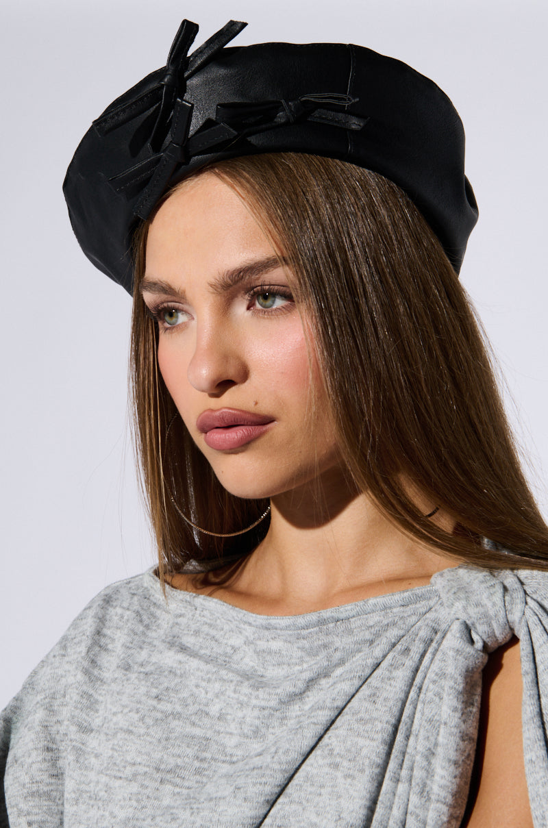 A person with long, straight hair wears a PRETTY IN PU BOW BERET and a gray top with tied shoulder detail. They have a neutral expression and are looking slightly to the side. The background is plain and light-colored. 1