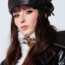 A person with long brown hair is wearing the stylish PRETTY IN PU BOW BERET in black pleather, paired with a black leather jacket featuring a white fur collar. With a confident expression and direct gaze into the camera, they exude a chic Parisian look against the plain white background.