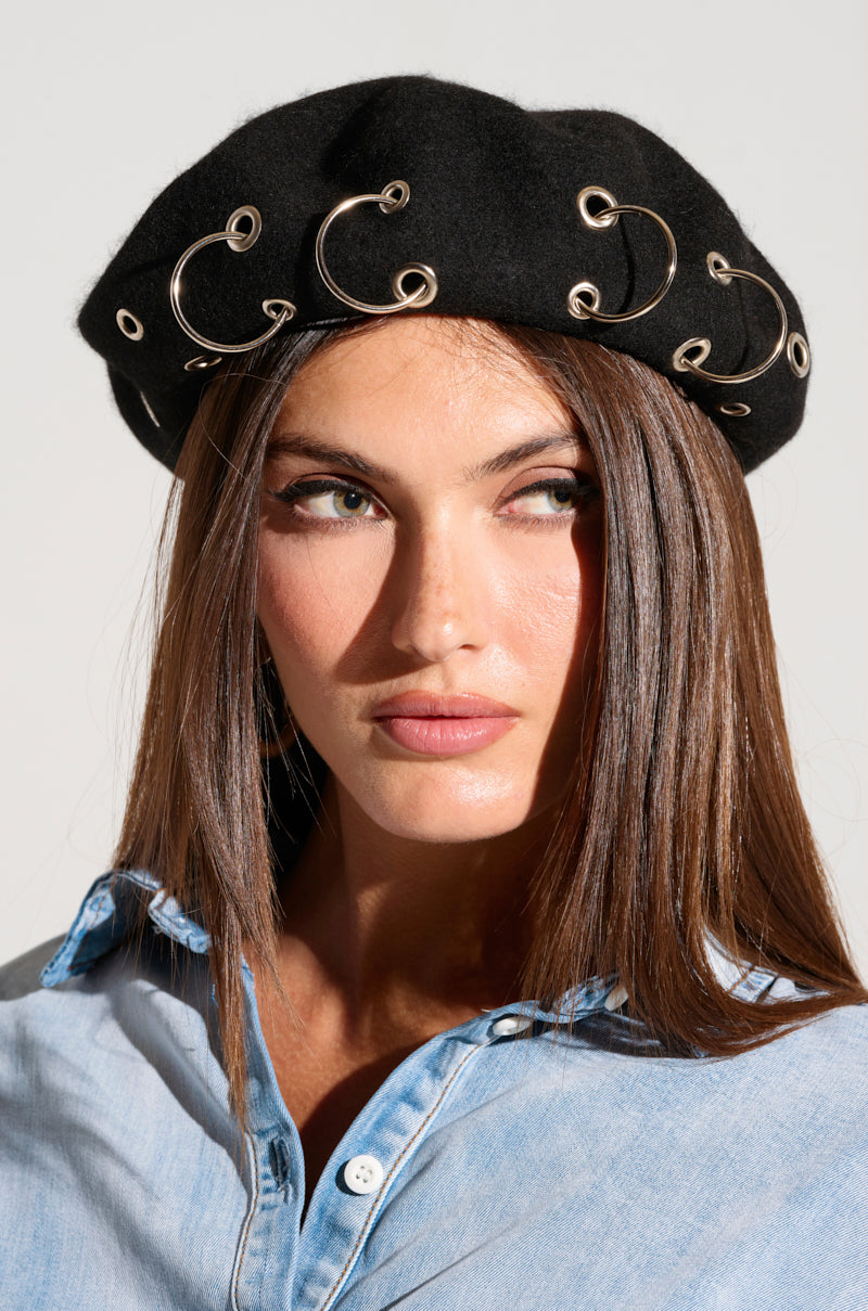 A woman with straight brown hair, wearing a light blue denim shirt and the STAY CONNECTED GROMMET BERET, which is decorated with multiple silver rings, gazes slightly to the right. She sports a natural makeup look with subtle blush and glossed lips against a plain, light-colored background.