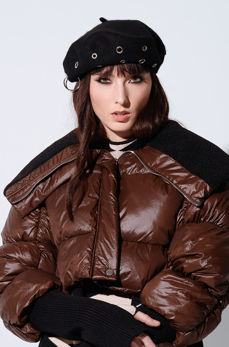 A person sporting a black STAY CONNECTED GROMMET BERET and a shiny brown puffer jacket with wide shoulders, perfect for fall fashion, is looking at the camera. The jacket has black accents, complementing the person's long, dark hair and fair skin.