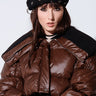 A person sporting a black STAY CONNECTED GROMMET BERET and a shiny brown puffer jacket with wide shoulders, perfect for fall fashion, is looking at the camera. The jacket has black accents, complementing the person's long, dark hair and fair skin.