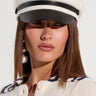 A person with long brown hair is wearing a TOO COOL BAKERS BOY CAP, which features a white body and faux leather black visor. They are dressed in a cream-colored textured top adorned with a navy pattern near the collar and sport an elaborate dangling pearl earring on the right ear. The background is plain and light.