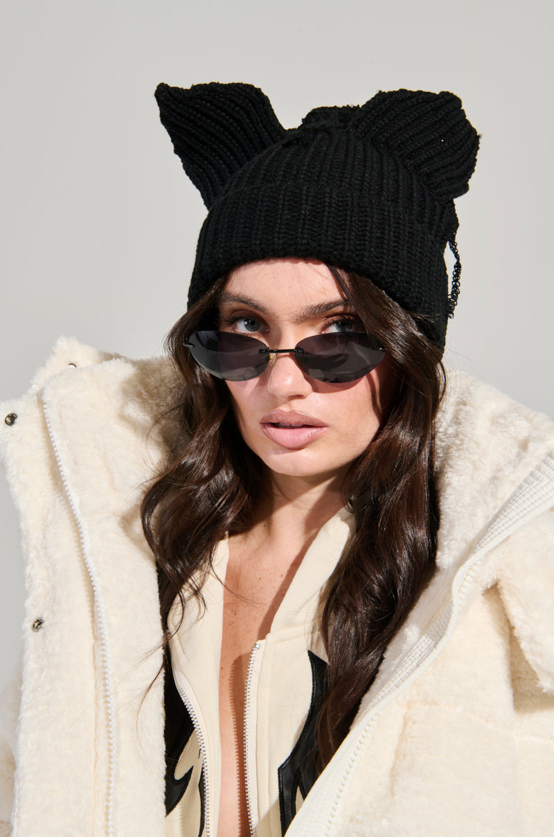 A person wearing a plush white winter coat and the IT GURL BEANIE stylishly pairs it with rectangular sunglasses. Their long, dark hair cascades gracefully, contrasting the neutral off-white background, and their top is partially unzipped.