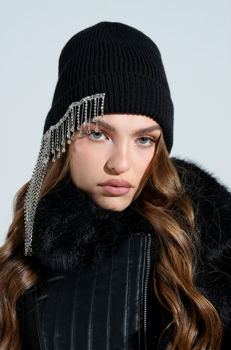 Sporting long, wavy hair under the RIRI RHINESTONE FRINGE BEANIE, a person pairs the blinged accessory with a black fur-lined jacket. Their winter style shines as they pose seriously against a light blue backdrop, radiating cool confidence.