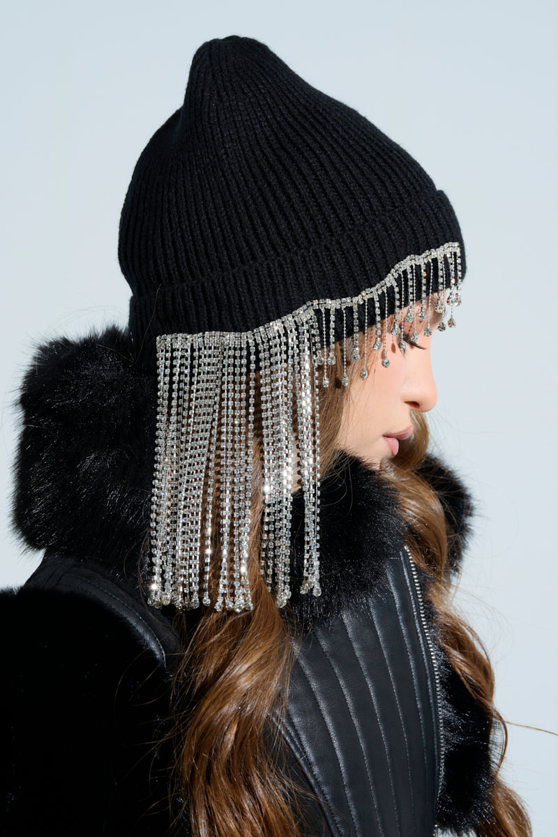Wearing the RIRI RHINESTONE FRINGE BEANIE, a person with long brown hair showcases winter style in a black fur-trimmed coat. The hat's sparkling rhinestone fringe drapes over their face, with a light blue background enhancing its allure.