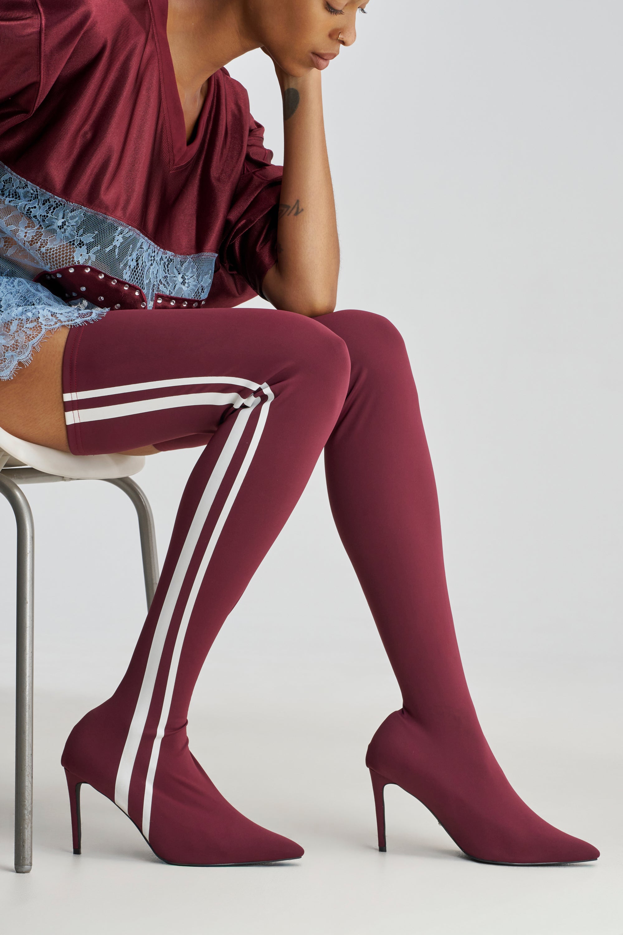 A person is seated on a chair, wearing a burgundy dress with blue lace trim and the AZALEA WANG THALUNE BURGUNDY STRETCH SPORTY STILETTO boots—burgundy thigh-high boots featuring two white stripes and pointed high heels. The image is cropped to display only the lower half of the body.