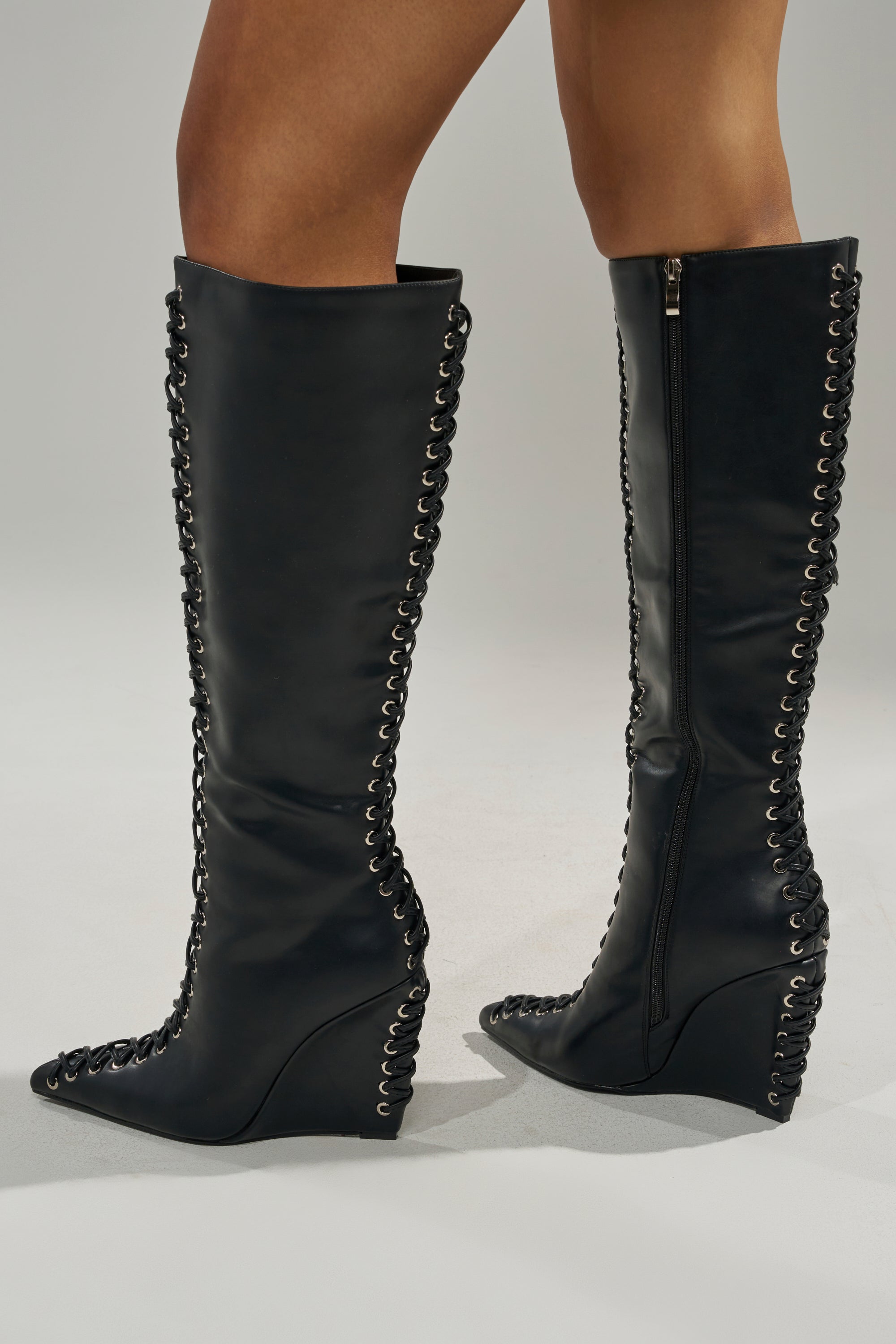 A person wears the AZALEA WANG THAUMUS BLACK LACE UP WEDGE BOOT, featuring silver eyelet details on the front and back, while standing on a light gray background. 1