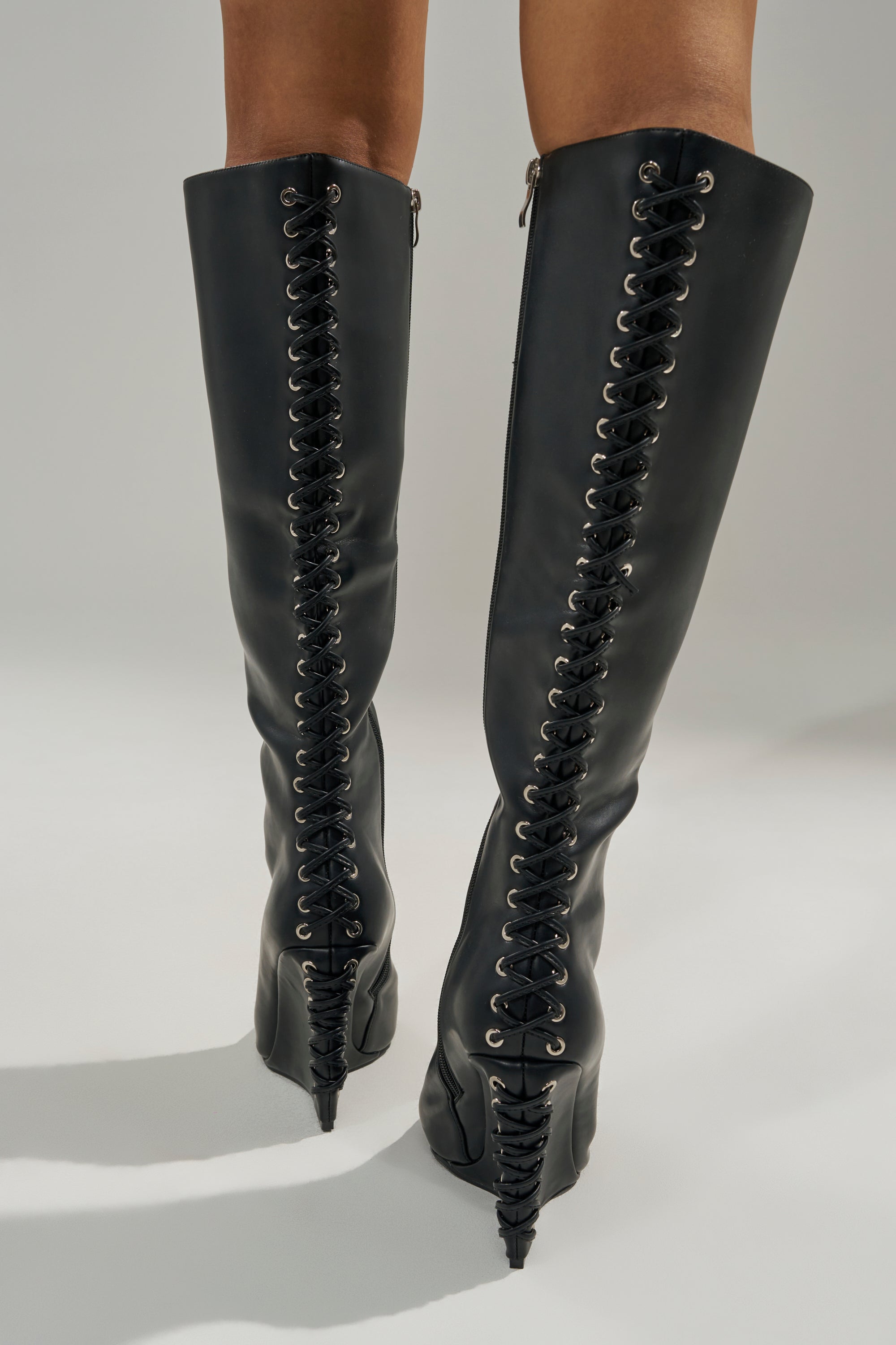 Close-up of a person wearing the AZALEA WANG THAUMUS BLACK LACE UP WEDGE BOOT, a faux leather knee-high with sculpted high heels, shown standing on a light grey surface. 3