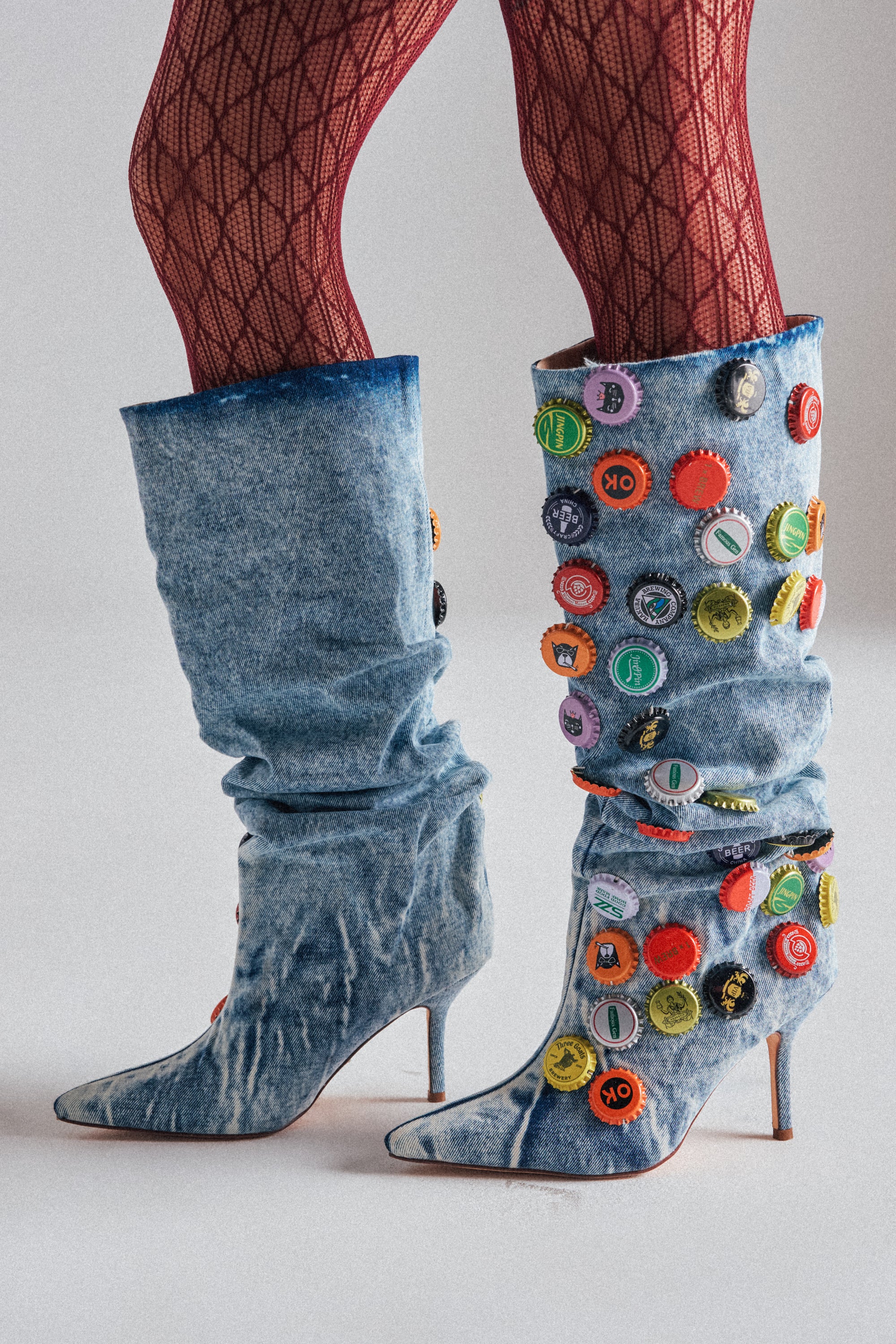 A person stands on a light background wearing AZALEA WANG THELEN DENIM BOTTLE CAP STILETTO BOOTS with bottle cap applique and blue denim, paired with red patterned tights. 0