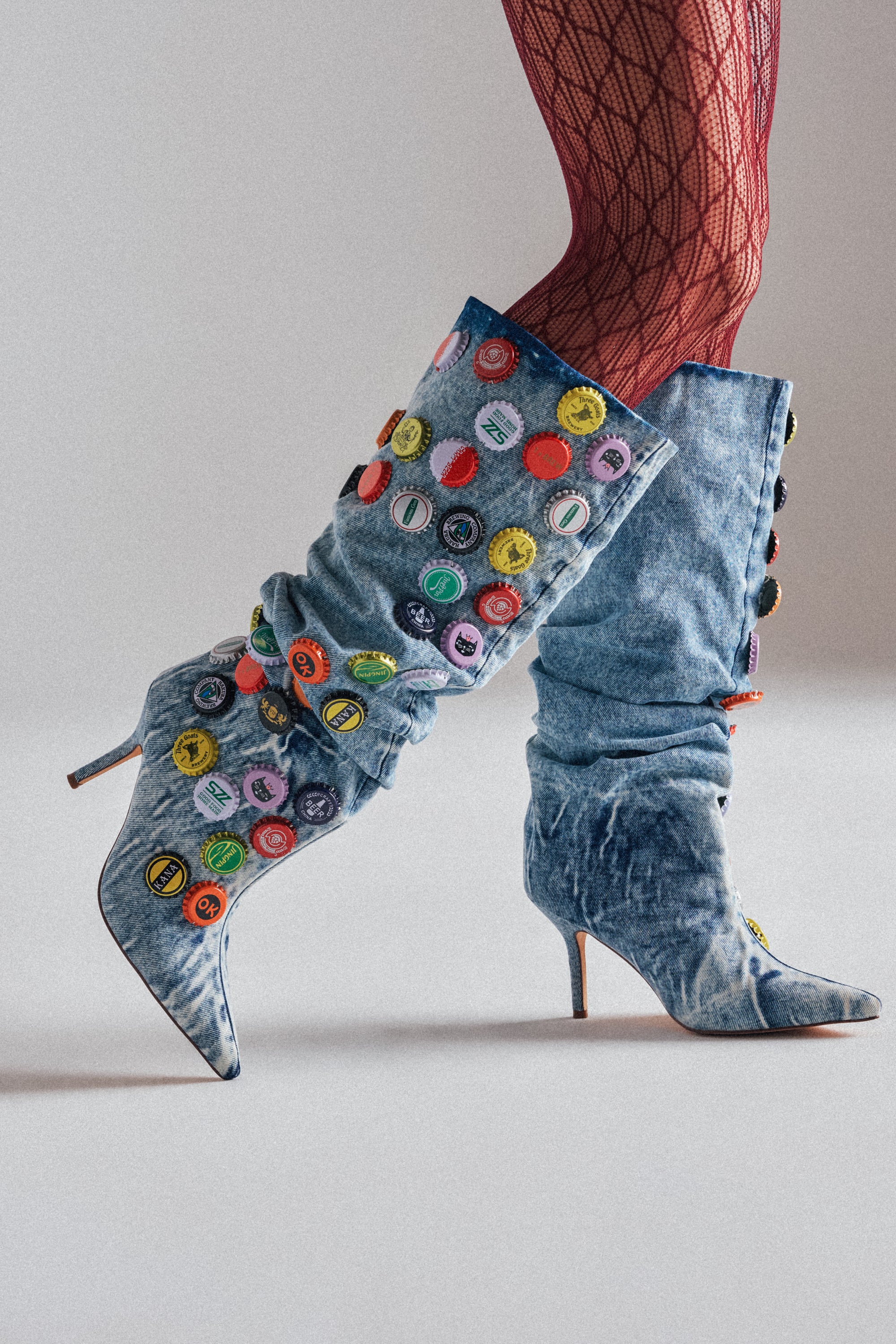 Model wears the AZALEA WANG THELEN DENIM BOTTLE CAP STILETTO BOOT in light blue with bottle cap appliqué and red fishnet stockings, posed against a plain light background. 1