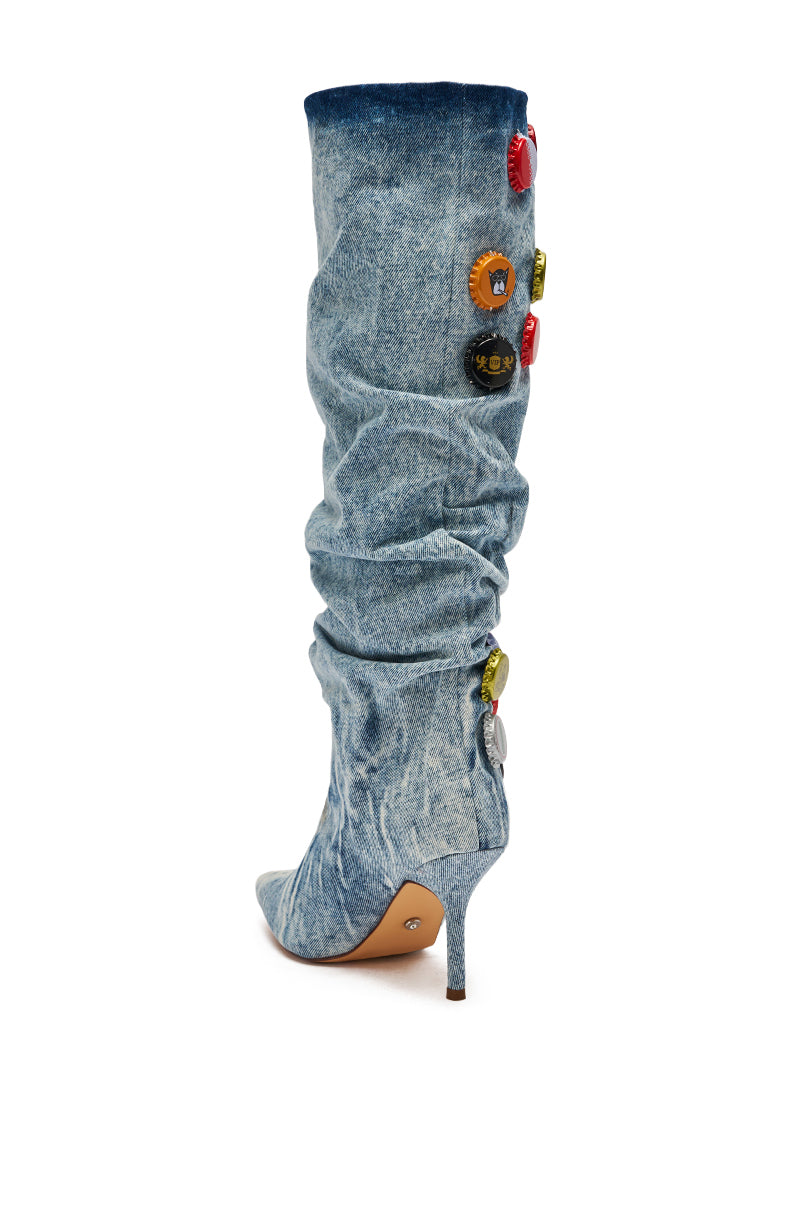 The AZALEA WANG THELEN DENIM BOTTLE CAP STILETTO BOOT is a knee-high, high-heeled style featuring a pointed toe, light wash denim, and bold bottle cap appliqué along the side and back.