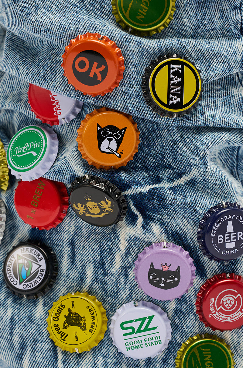 Colorful bottle caps with cat and text logos are scattered on blue denim, capturing the playful vibe of the AZALEA WANG THELEN DENIM BOTTLE CAP STILETTO BOOT.