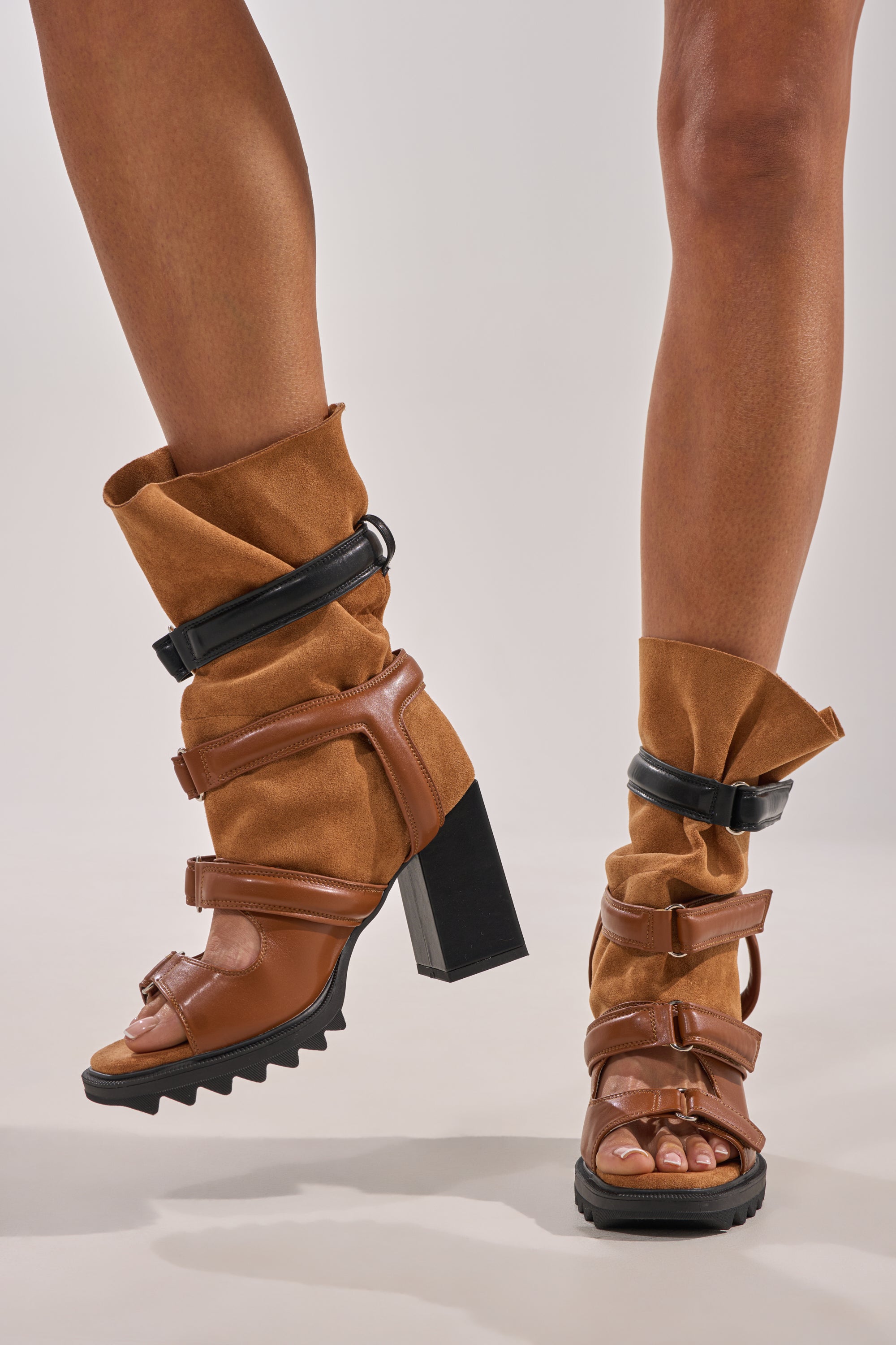 A pair of legs wearing the AZALEA WANG THIERRY Tan Faux Suede Open Toe Chunky Bootie with thick, rugged soles and multiple straps, paired with slouchy, tan faux suede leg warmers reaching mid-calf. 0