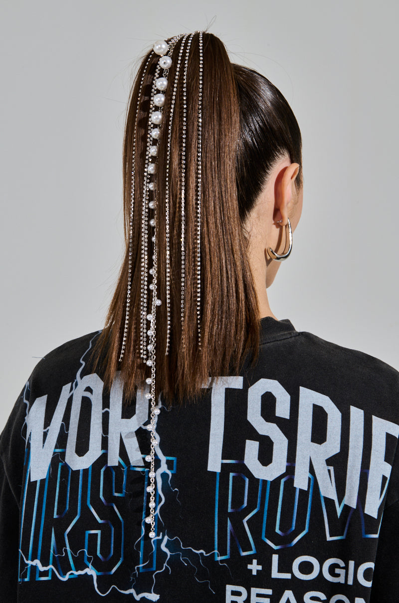 The individual sports long brown hair in a high ponytail, accessorized with the DRIPP DIAMONDS HAIR TIE featuring chains and beads with pearl accents. They wear a black graphic t-shirt with light blue and white text, large hoop earrings, all set against a plain gray background.