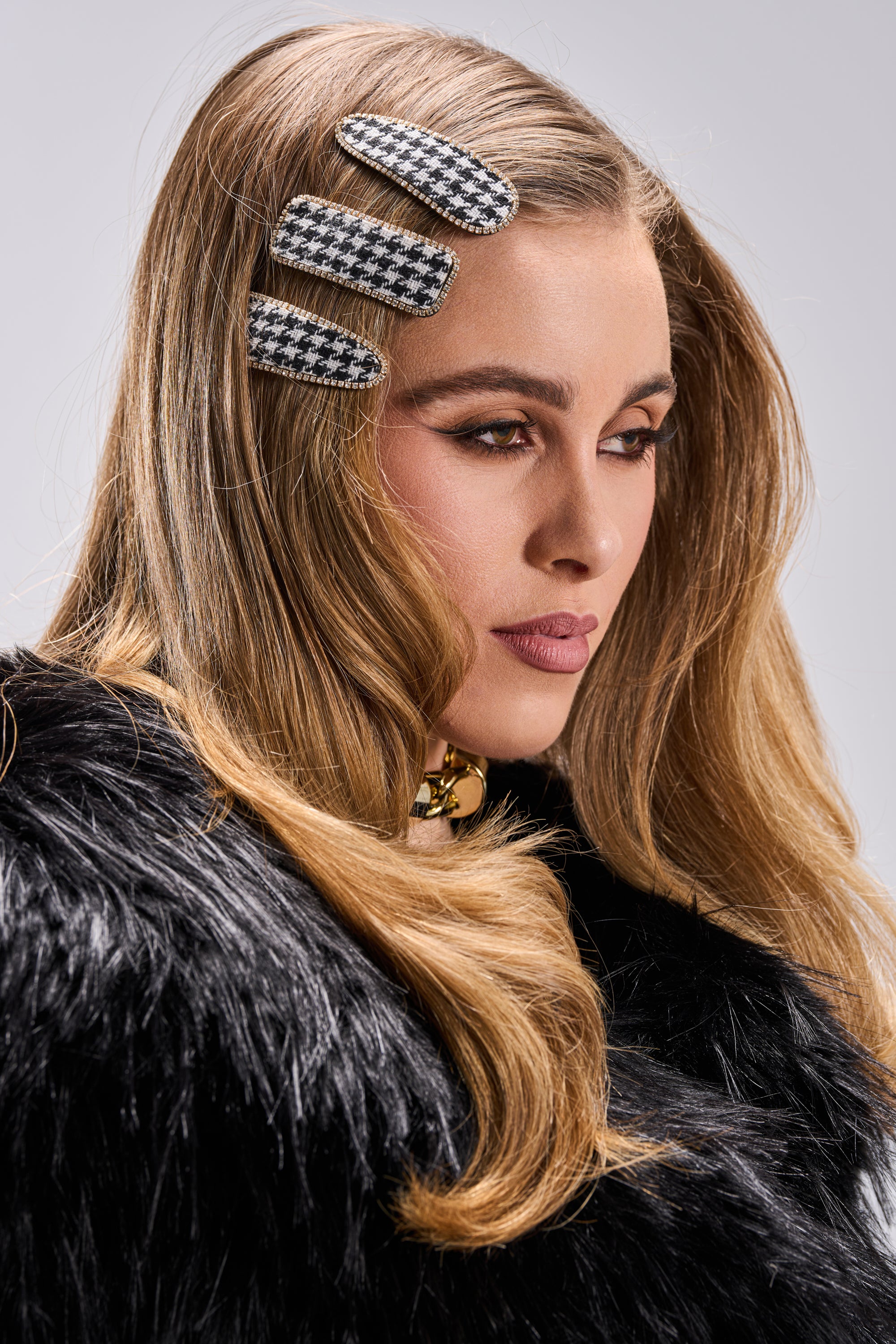 A woman with long, wavy blonde hair styles her look with three large COCO HAIR CLIPS and a black faux fur coat, finishing her ensemble with a chunky gold necklace and a serious expression.