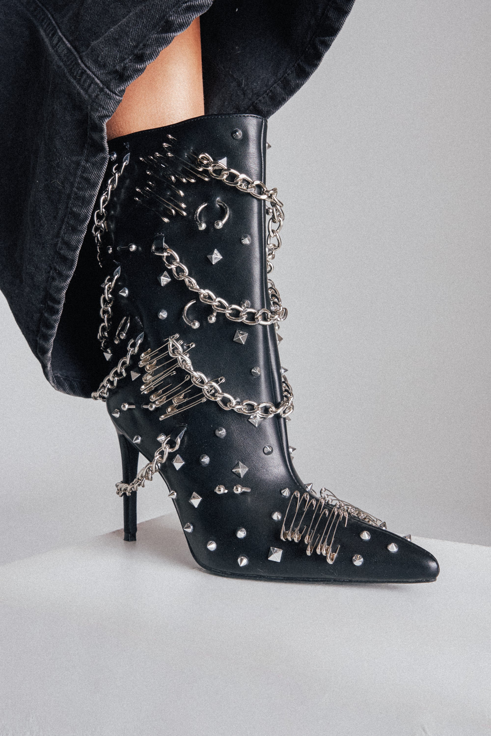 The AZALEA WANG THRIGRUN BLACK SILVER HARDWARE STILETTO BOOTIE is a faux leather, pointed toe bootie adorned with silver spikes, studs, safety pins, and chains for a bold punk look—styled here with black pants.