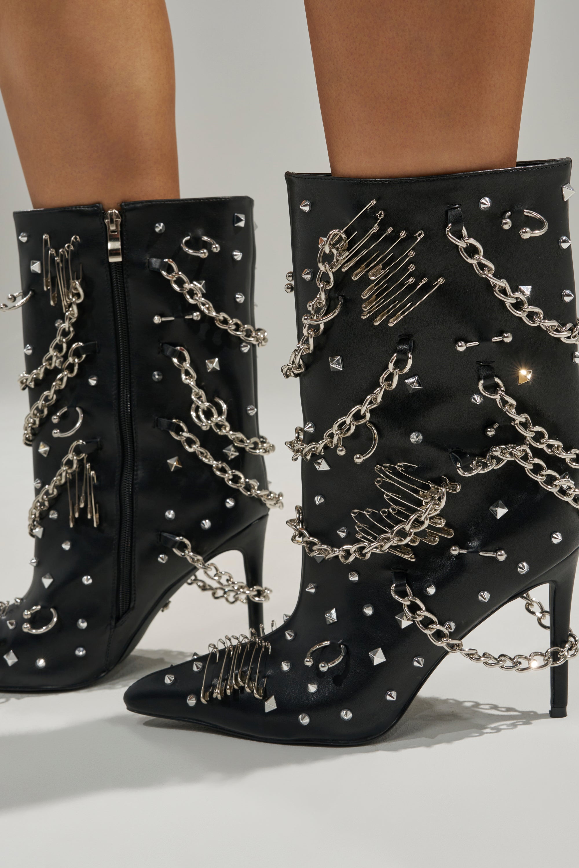The AZALEA WANG THRIGRUN BLACK SILVER HARDWARE STILETTO BOOTIE features black faux leather, pointed toes, side zippers, and edgy silver chains, safety pins, and studs—styled here on bare legs.