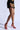 A person stands in profile wearing a black dress adorned with gold and red heart designs, complemented by striking AZALEA WANG THUNDERMINE Red Velvet Embellished Stiletto Sandals. The background is a light neutral shade, enhancing the elegance of the ensemble.