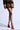 A person is dressed in a black dress adorned with gold and red heart patterns, complete with a front zipper. They are posed against a light background, drawing attention to their footwear—the AZALEA WANG THUNDERMINE RED VELVET EMBELLISHED STILETTO SANDALS—with the image focusing on the lower half of the body to emphasize this chic choice.