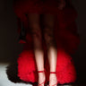 In dramatic lighting, someone wears a voluminous red ruffled gown and AZALEA WANG THUNDERMINE red velvet embellished stiletto sandals with comfort cushions. The gown's asymmetric hem showcases their legs while holding a black studded clutch.