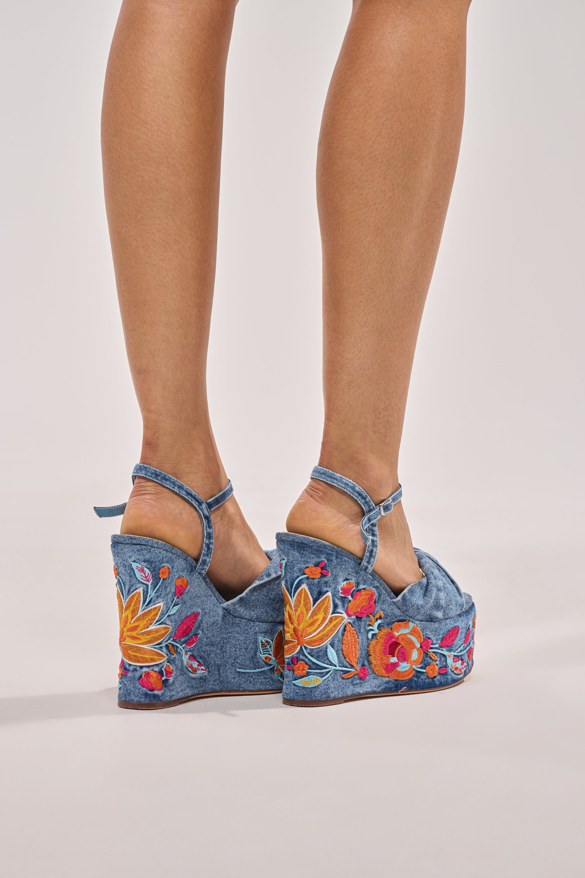 A person wears the AZALEA WANG TIALA DENIM FLORAL EMBROIDERED STRAP WEDGE SANDAL with ankle straps, platform heels, and colorful floral embroidery, standing on a light background. 3