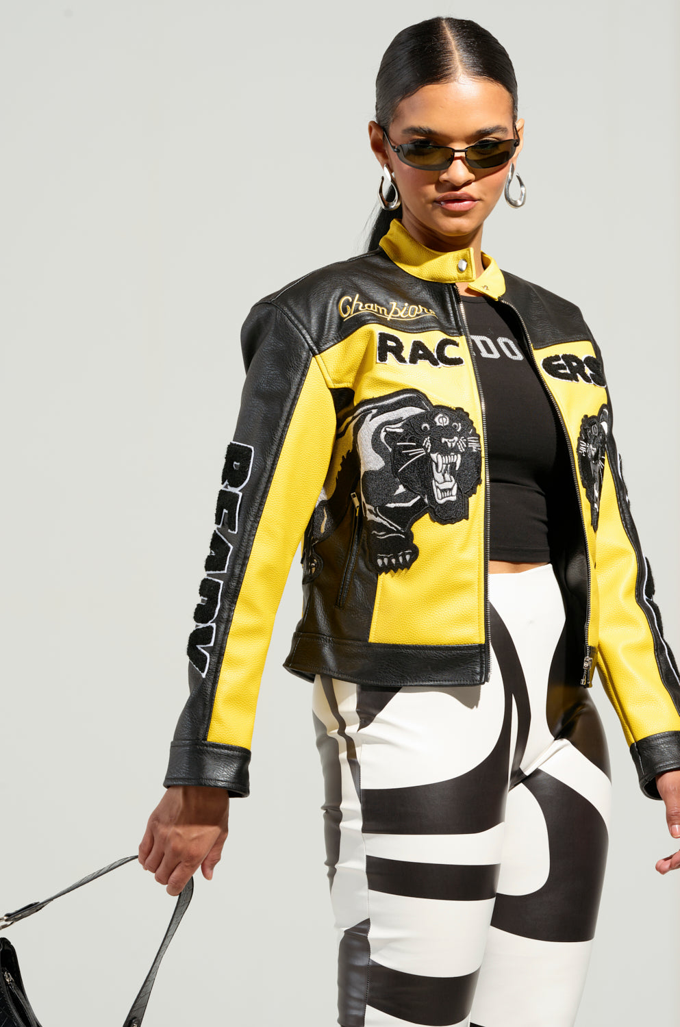 A person confidently stands against a neutral background, wearing the EYE OF THE TIGER MOTO JACKET—an eye-catching yellow and black faux leather piece featuring racing-themed patches and tiger patch detailing on its color block paneling. Complementing the jacket with bold black and white pants and sunglasses, they hold a handbag with style.