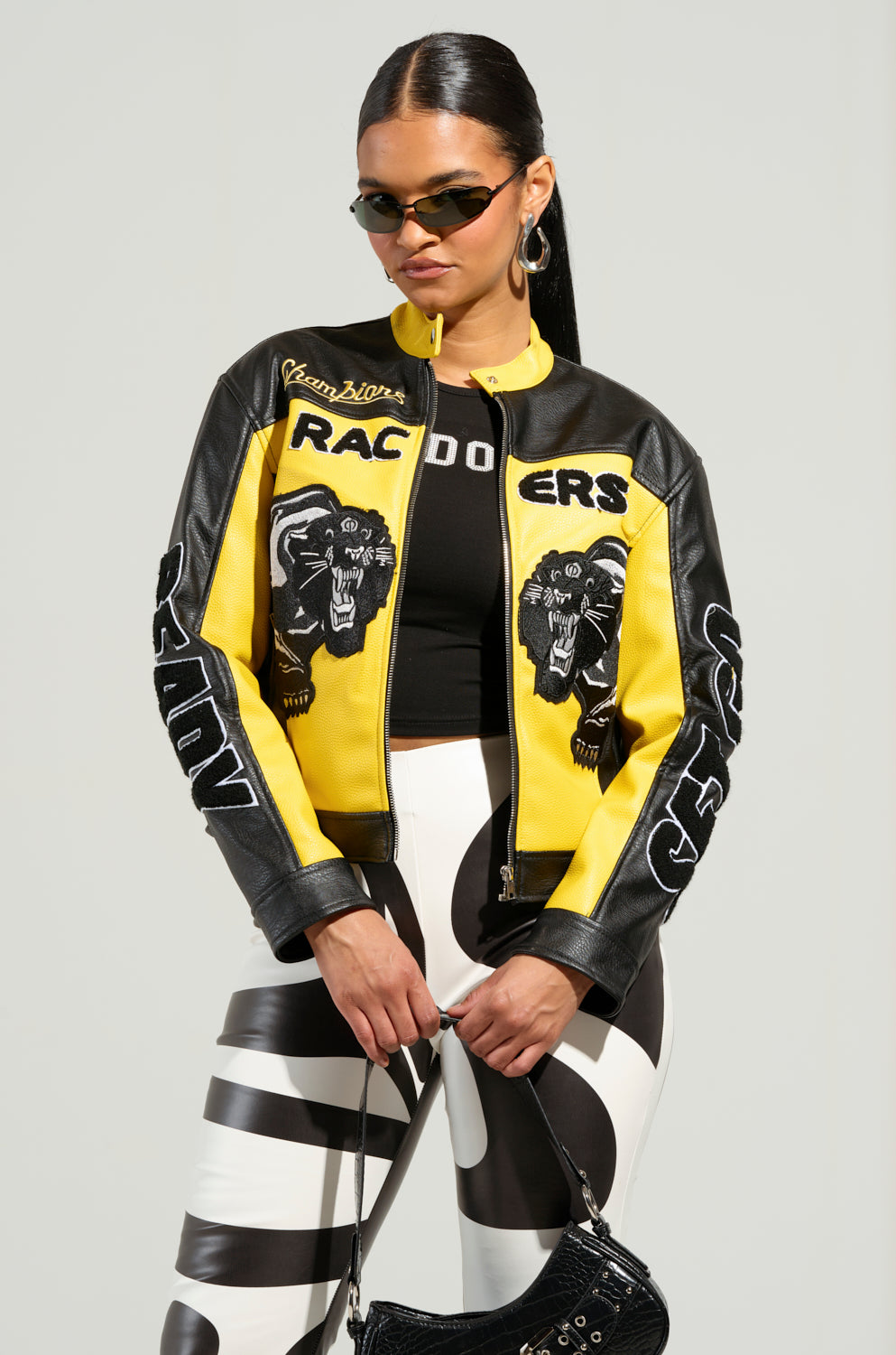 The woman confidently rocks the EYE OF THE TIGER MOTO JACKET, featuring striking black and yellow faux leather with bold "Racers" and panther designs. Her look is completed with a black crop top, color block paneling pants, sunglasses, hoop earrings, and a sleek black purse. Her hair is pulled back smoothly for an effortlessly chic appearance.