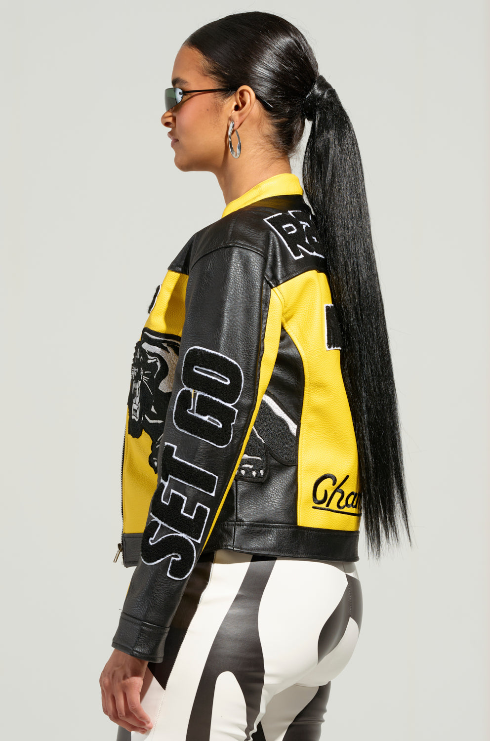 A person with long black hair styled in a sleek ponytail is wearing sunglasses and the EYE OF THE TIGER MOTO JACKET, featuring yellow and black color block paneling and tiger patch detailing. They pair it with black and white patterned pants, standing against a plain background.