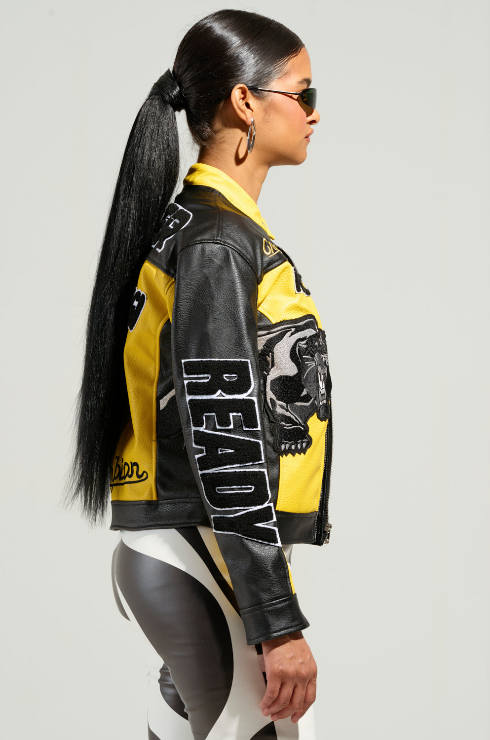 A woman with long hair in a ponytail is wearing the *EYE OF THE TIGER MOTO JACKET*, black sunglasses, and patterned pants. She stands in profile against a plain background.