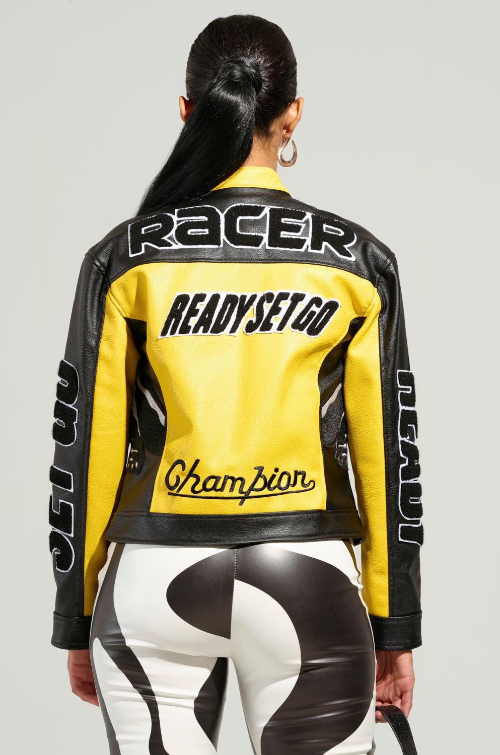 A person with long hair stands facing away, wearing the EYE OF THE TIGER MOTO JACKET, a yellow and black faux leather jacket featuring the words "RACER" and "READY SET GO" on the back. The jacket showcases color block paneling and is paired with black and white patterned pants.