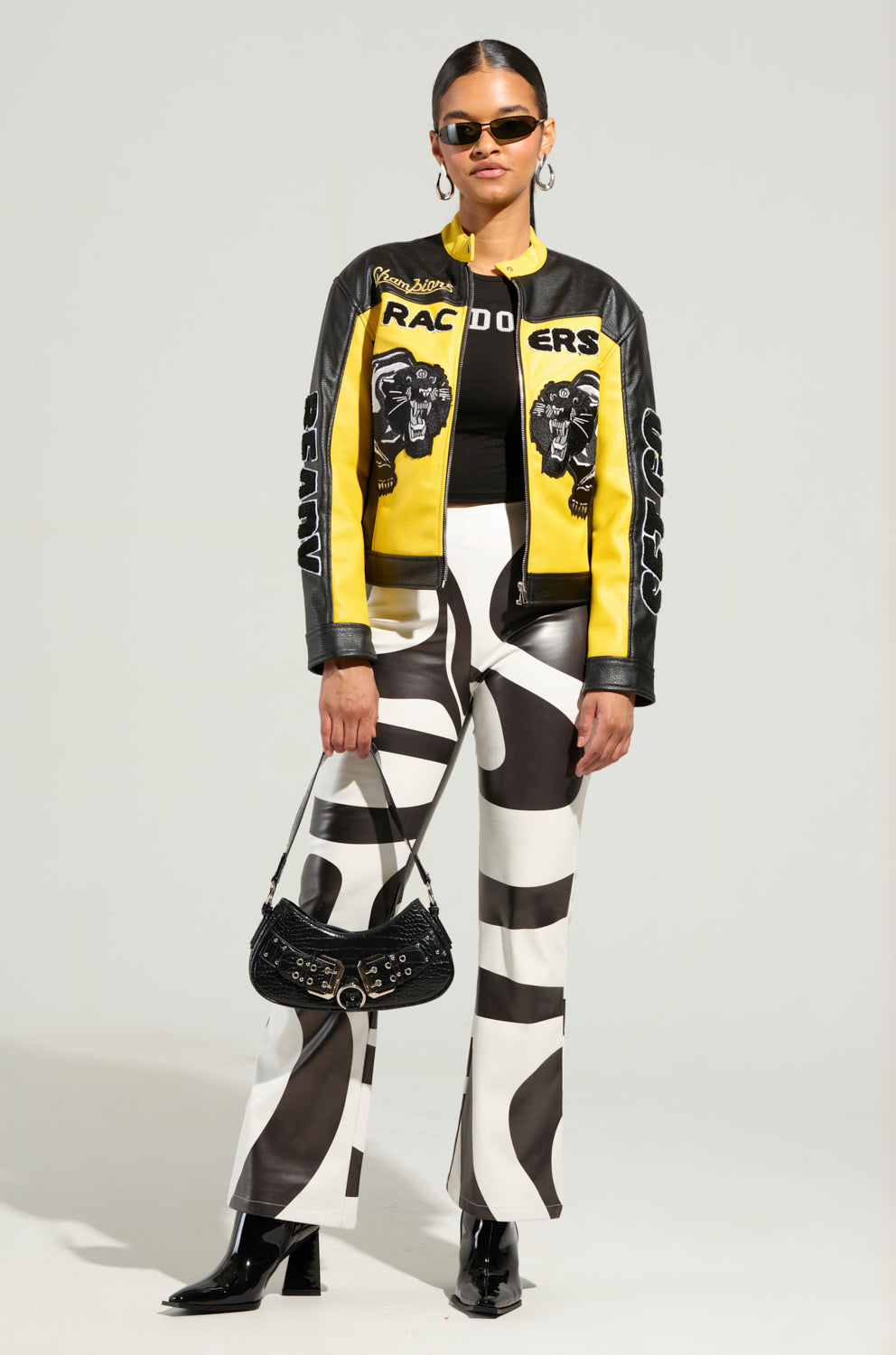 A person sports the EYE OF THE TIGER MOTO JACKET in yellow and black with "Racers" text, graphic patterns, and tiger patch detailing. They pair it with a black top, bold black-and-white patterned pants, black boots, sunglasses, and a chic black handbag against a plain backdrop.