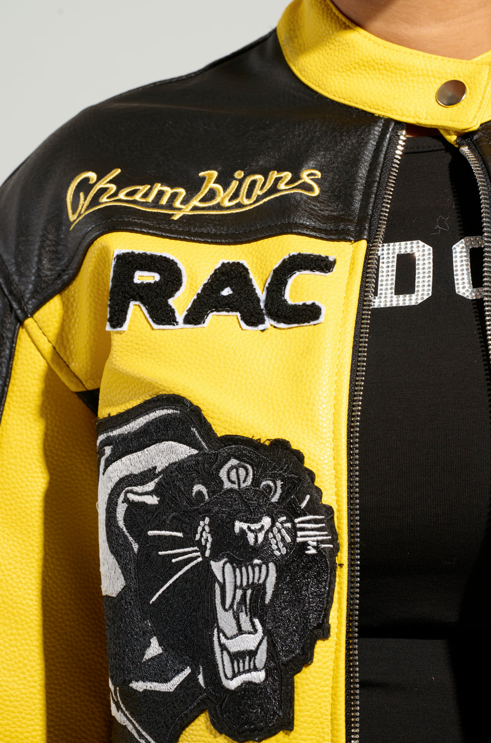 Close-up of the EYE OF THE TIGER MOTO JACKET in yellow and black faux leather, showcasing "Champions RAC" wording with a tiger patch on the sleeve. A glimpse of a black shirt underneath accentuates the jacket's bold color block design.