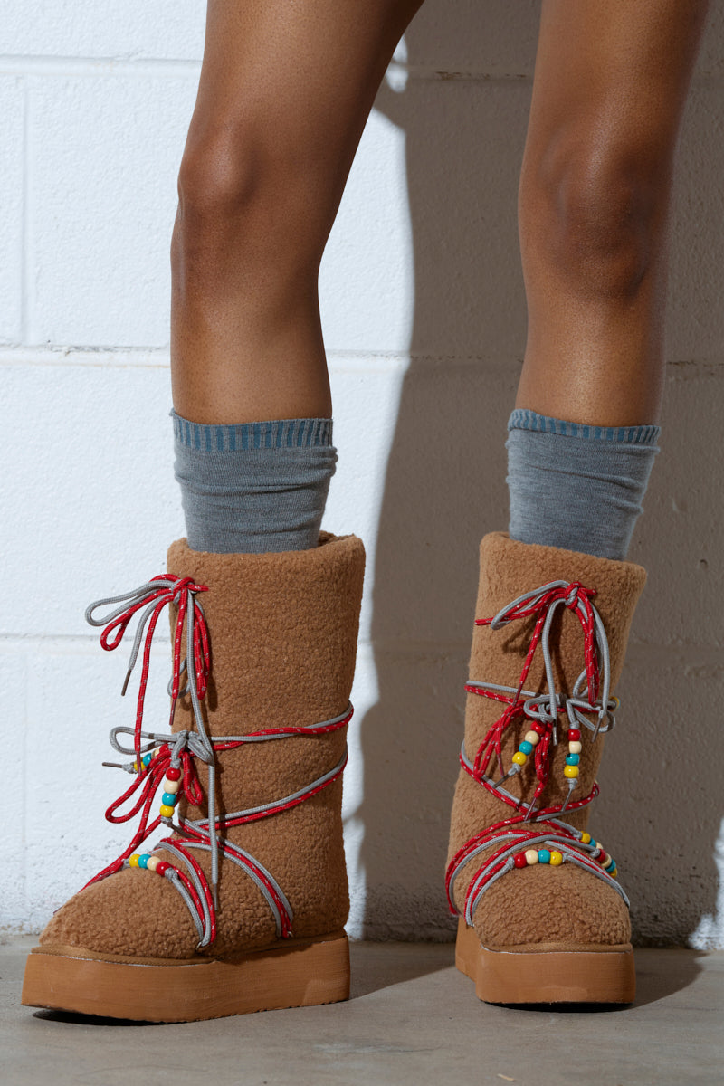 A person stands against a light brick wall wearing the TIGHTROPES CHESTNUT SHERPA WITH BEADS FLAT BOOTIE. The booties have a faux sherpa upper, a flatform sole, gray socks underneath, and colorful red, gray, and white laces adorned with beads for extra style.