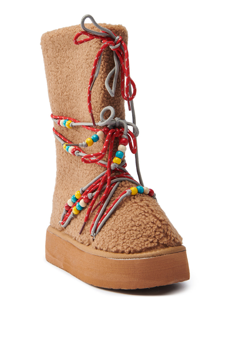 The TIGHTROPES CHESNUT SHERPA WITH BEADS FLAT BOOTIE features a tan, fleece-textured faux Sherpa upper and a thick flatform sole, adorned with red and gray laces with colorful beads. Its plush look makes it ideal for cold weather.