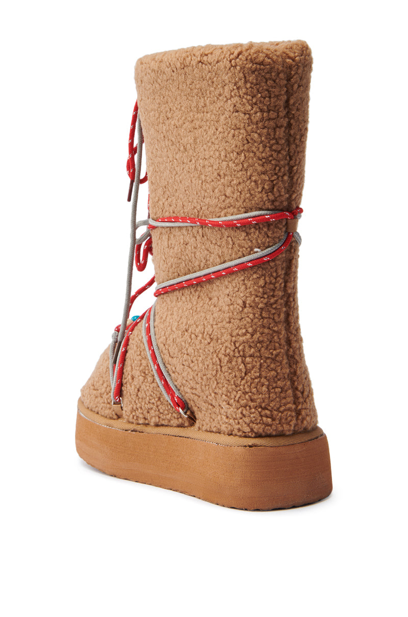 The TIGHTROPES CHESNUT SHERPA WITH BEADS FLAT BOOTIE features a mid-calf faux Sherpa chestnut upper, a rubber sole, and stylish red and gray laces wrapped around it for a cozy look.