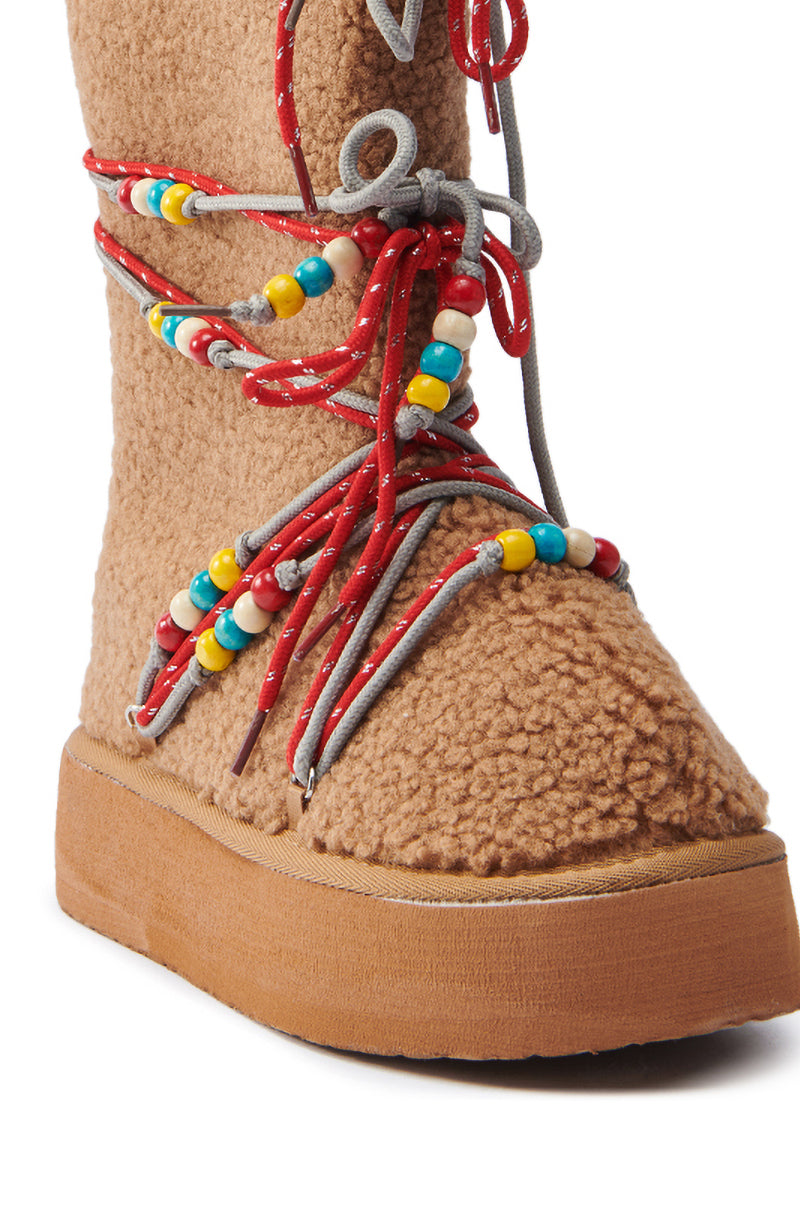 The TIGHTROPES CHESNUT SHERPA WITH BEADS FLAT BOOTIE features a tan faux Sherpa upper and thick flatform sole, highlighted by colorful beads adorning red and gray laces crisscrossing up the front.