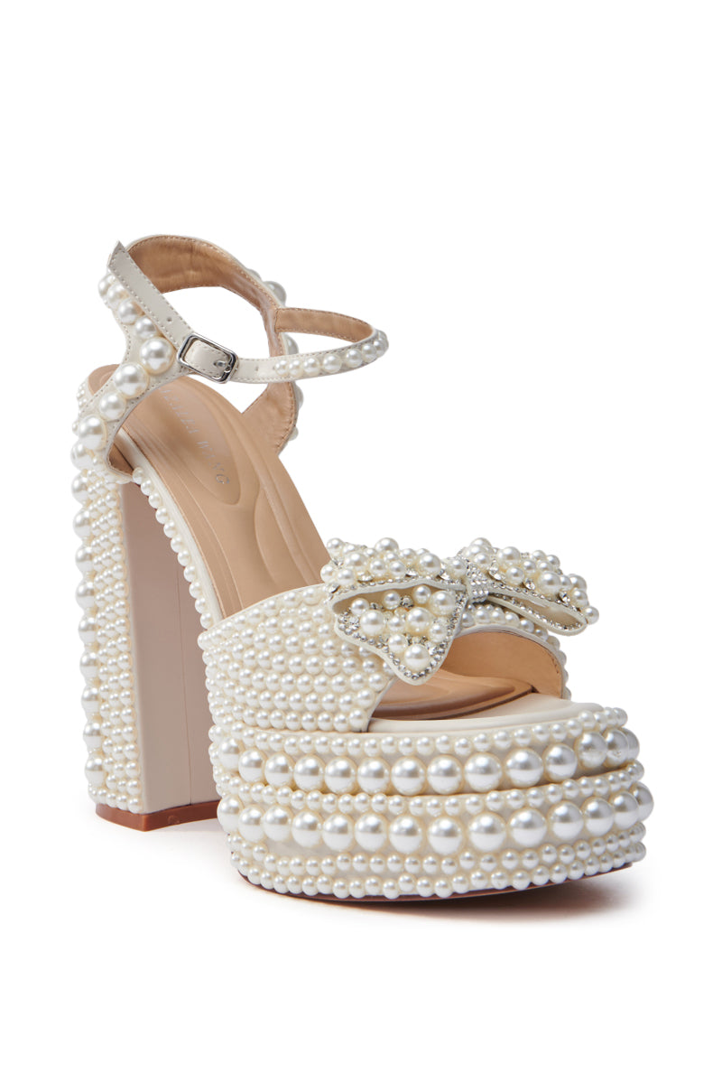 The AZALEA WANG TIMEA WHITE CHUNKY SANDAL is a high-heeled shoe with a mega-chunky heel and ankle strap, embellished with white pearls. It features a decorative bow on the front, also adorned with pearls, and includes a comfort cushion insole for added ease.
