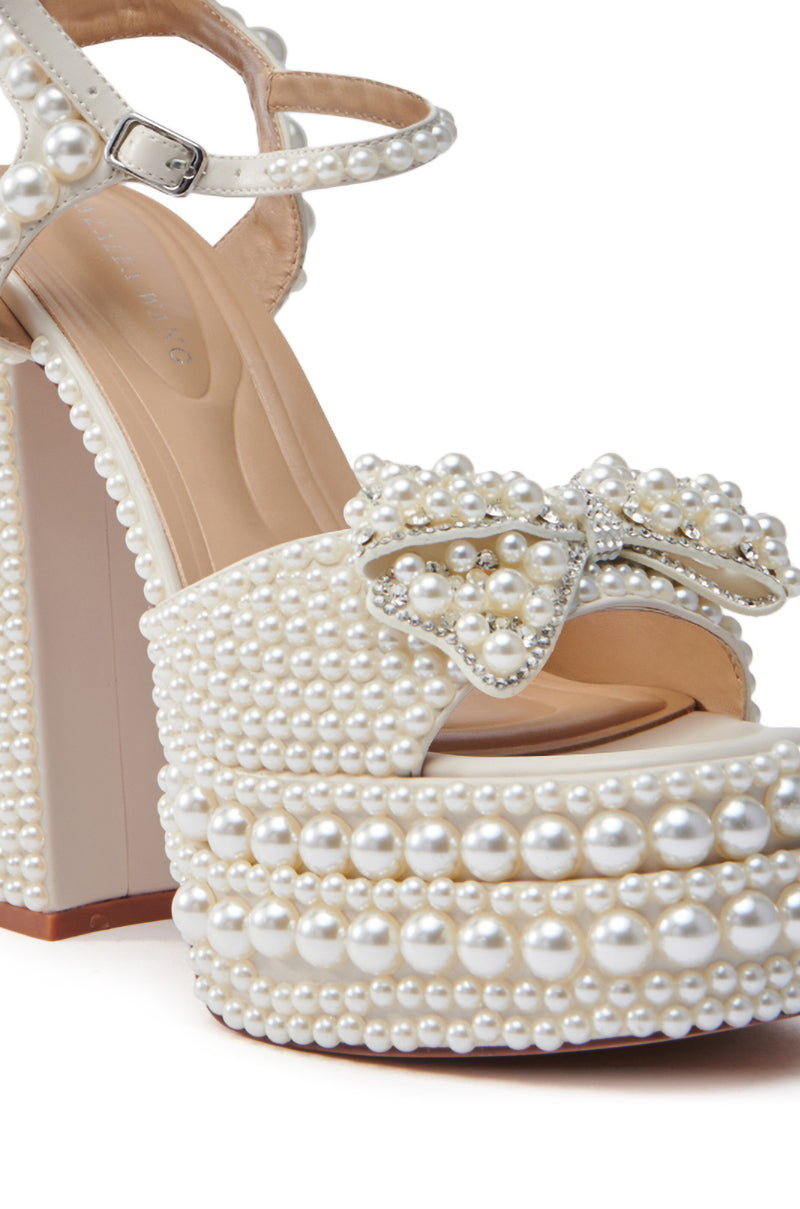 The AZALEA WANG TIMEA WHITE CHUNKY SANDAL is a high-heeled platform featuring white pearls, an open-toe design with a pearl bow on the front strap, and a comfort cushion insole. It includes an ankle strap with a silver buckle for added style.
